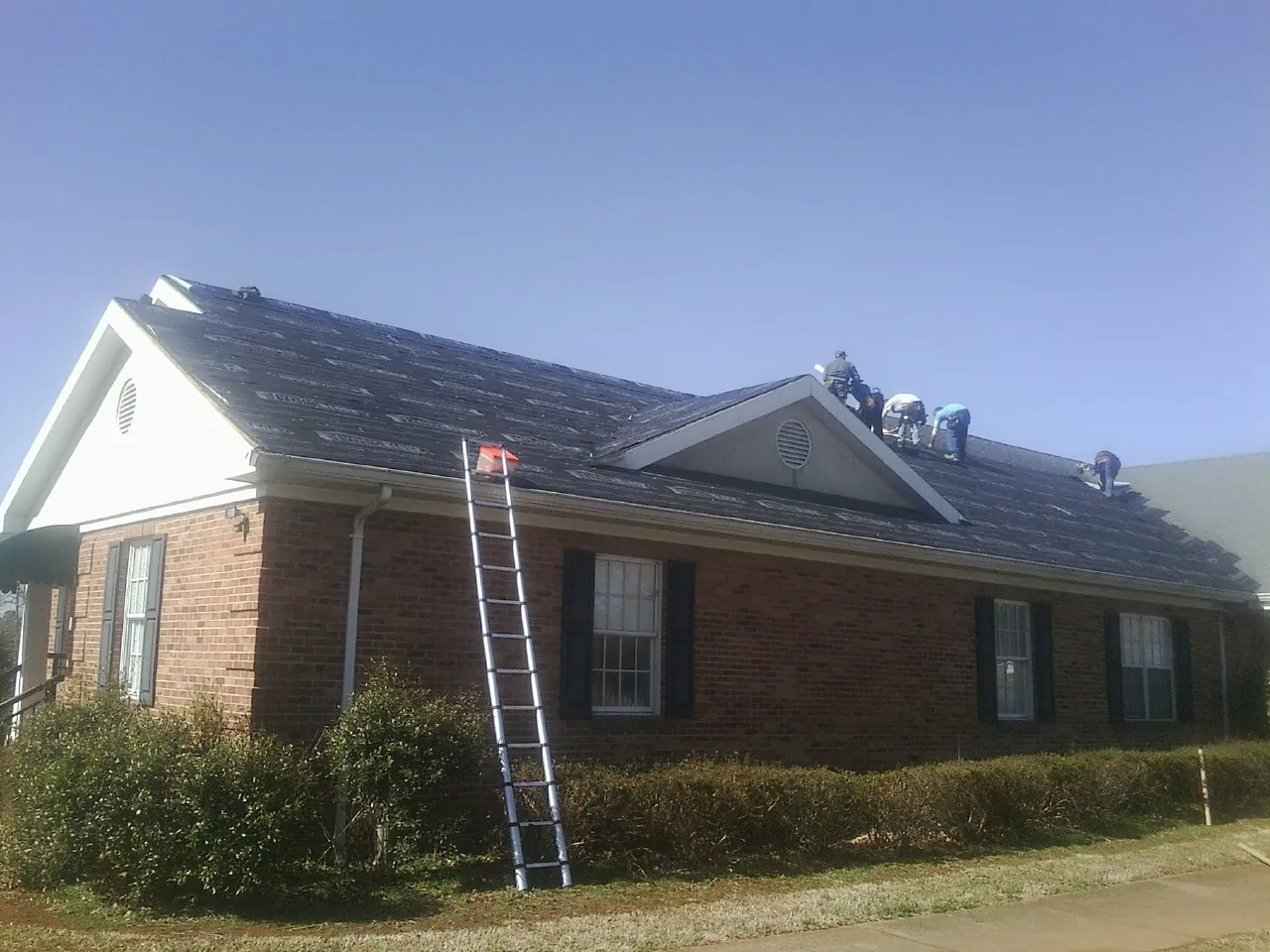 Crew tearing shingles off a brick ranch roof with a ladder set — Metal Roof Repair work by our crew in Kinston