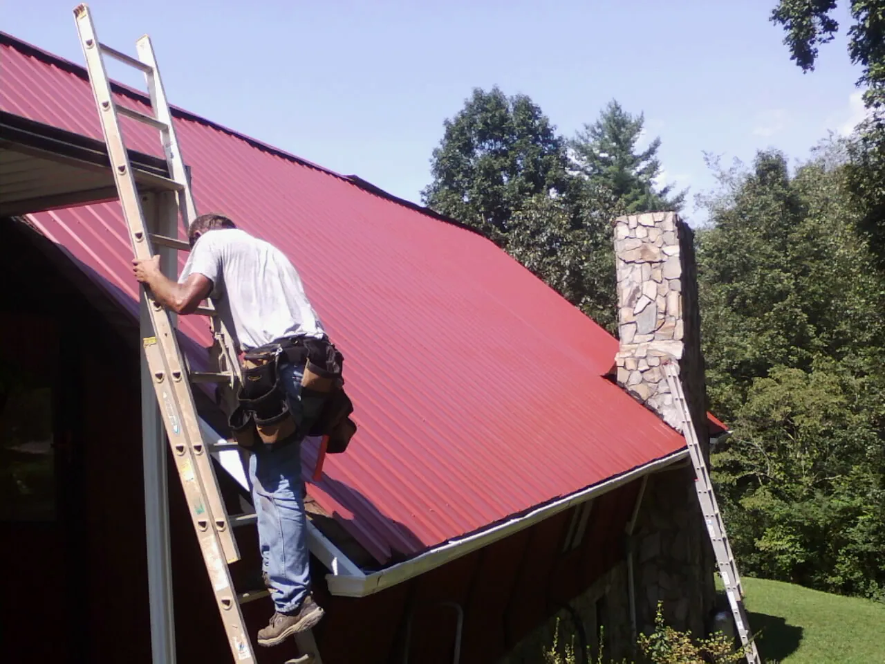Roofer on a ladder beside a red metal roof and stone chimney — Soffit & Fascia Repair by Roofing Brothers in Kinston